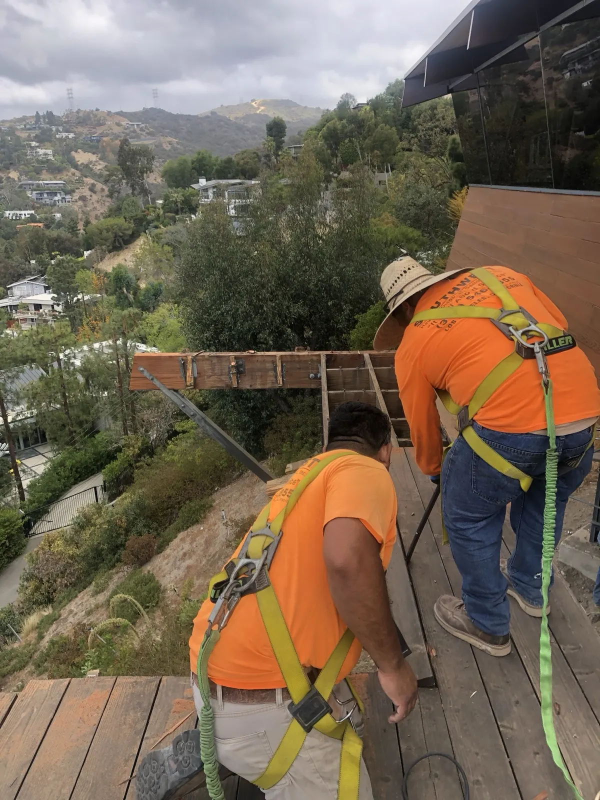 Two crew members in safety harnesses taking apart a deck on a hillside above Los Angeles